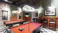 This dedicated billiards lounge offers a well-appointed recreational space designed for serious pool enthusiasts and casual players alike. The centerpiece of the room is two full-size pool tables dressed in bold red felt, each illuminated by ornate stained-glass pendant lights that lend a timeless, tavern-inspired aesthetic. The flooring features a striking black-and-white checkered tile pattern that anchors the room's classic character. Deep green walls are complemented by warm wood wainscoting and exposed stone brick accents, creating an inviting, pub-like atmosphere. A wall-mounted cue rack keeps equipment neatly organized, while framed artwork and a decorative ball display case add to the room's thematic charm. A flat-screen television is mounted on the wall, offering entertainment beyond the tables. The bar area is outfitted with tall bistro-style tables and black bar stools, providing a comfortable spot for guests to gather and relax. The exposed ceiling structure adds an industrial touch that contrasts nicely with the warm, traditional finishes throughout. Overall, the space strikes a balance between a functional game room and a stylish social lounge.