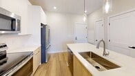 This 930-square-foot, single-floor apartment at 2418 Main St in Rocky Hill, CT offers a well-designed layout with 2 bedrooms, 2 bathrooms, 2 closets, and a hallway. The open-concept kitchen and living room serve as the heart of the unit, featuring light wood-look flooring, warm neutral walls, and French doors that invite natural light. The kitchen is outfitted with modern white upper cabinetry, natural wood-toned lower cabinets, a white quartz countertop island with a stainless steel undermount sink, pendant lighting, and a full suite of stainless steel appliances including a French door refrigerator, electric range, and over-the-range microwave. The living area includes a ceiling fan and direct outdoor access through glass-paneled doors. Both bedrooms are carpeted and include ceiling fans; the secondary bedroom features a large window with outdoor views. The primary bedroom, at 140 square feet, connects to a bathroom with a wood-toned vanity, white quartz countertop, and a tub-shower combo. Each bedroom is accompanied by a walk-in closet with wire shelving systems. Clean lines, neutral tones, and contemporary finishes throughout give this apartment a fresh, modern ambiance.