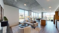 This single-floor residence showcases a distinctly urban aesthetic defined by exposed concrete ceilings, warm hardwood flooring, and expansive floor-to-ceiling glass walls that flood the interior with natural light and frame sweeping city views. The open-plan living and dining area is anchored by a generous seating arrangement and flows seamlessly toward a corner glazed dining nook, where exposed concrete columns punctuate the glass perimeter—creating an industrial-modern focal point ideal for daily living and entertaining.

The kitchen is outfitted with warm wood cabinetry, light stone countertops, a marble-style tile backsplash, stainless steel appliances including a gas range, and a deep undermount sink. A central island with wood-paneled detailing provides additional workspace and casual seating.

The primary bedroom features the same exposed concrete ceiling treatment, a four-poster bed frame, and a spacious layout with curated wall art. A second bedroom offers comparable comfort with a barn-style sliding door and ceiling fan. Both bathrooms are finished with wood-toned vanities, light stone surfaces, and hardwood-style flooring. A dedicated laundry area houses a full-size side-by-side washer and dryer. A compact powder room completes the layout.