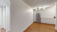 This single-floor space offers a clean, minimalist aesthetic defined by bright white walls, a textured popcorn ceiling, and warm parquet wood flooring that flows consistently throughout the unit. The open layout connects the living area to the kitchen, which is equipped with stainless steel appliances including a full-size refrigerator and an electric range, complemented by ample white cabinetry and generous counter space along two walls. A distinctive decorative partition with open shelving separates the kitchen from the living area, adding a subtle architectural detail to the otherwise streamlined interior. Track lighting illuminates both the kitchen and living spaces, providing functional and well-distributed light. The spacious main room benefits from a large window fitted with horizontal blinds, allowing natural light to fill the space and highlight the polished parquet floors. The bathroom maintains the unit's clean aesthetic with fully white-tiled walls, a walk-in shower with a safety grab bar, and a window for added ventilation and natural light. Overall, the space presents a practical and cohesive design that emphasizes functionality, bright open areas, and consistent finishes throughout.