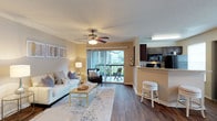 This single-story residence features an open-concept design that seamlessly connects the living area and kitchen, creating a cohesive and functional layout. The living room is anchored by warm hardwood-style flooring that extends throughout the space, complemented by neutral beige walls that provide a welcoming ambiance. A ceiling fan with integrated lighting serves as a central fixture, enhancing both comfort and airflow. Large sliding glass doors flood the room with natural light and open directly onto a private balcony, where outdoor seating allows for alfresco relaxation with views of surrounding greenery.

Adjacent to the living area, the kitchen is defined by rich, dark wood cabinetry and a granite-style countertop that wraps into a breakfast bar with barstool seating — ideal for casual dining. Black appliances, including a built-in microwave and range, provide a sleek, uniform aesthetic. The textured ceiling adds subtle dimension overhead, while the open sightlines between the kitchen and living room ensure the space feels connected and airy throughout.
