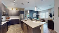 This single-floor residence offers a well-designed open-concept layout connecting the kitchen and living areas. The kitchen is equipped with dark shaker-style cabinetry, stainless steel appliances including a refrigerator, range, and microwave, complemented by light quartz countertops and wood-look flooring. A central island with an undermount sink and pendant lighting serves as both a functional workspace and casual dining area. Track lighting above adds further illumination to the space.

The living area features multiple windows that invite natural light, a ceiling fan, and a bold navy accent wall that anchors the room. The two bedrooms offer plush carpeting, large windows with exterior views, and ample space for furnishings. One bedroom connects directly to a full bathroom with a soaking tub, shower, and wood-look tile flooring, with an adjacent walk-in closet visible beyond.

A second full bathroom also features a tub-shower combination. A dedicated utility and pantry room houses a top-load washer alongside generous built-in open shelving, providing practical storage. Throughout the residence, a cohesive neutral palette with modern finishes creates a clean, contemporary ambiance.