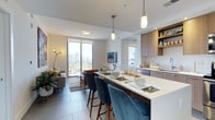 This single-floor space showcases a bright, open-concept layout that unifies the kitchen, dining, and living areas into one cohesive environment. The kitchen is anchored by a large island with a crisp white countertop, offering seating for multiple guests via plush teal upholstered bar stools. Warm wood-toned cabinetry lines the kitchen walls, complemented by open shelving that adds both storage and visual depth. Pendant lighting with an industrial-modern aesthetic hangs above the island, adding a stylish focal point. The adjacent dining and living area flows naturally from the kitchen, featuring light-toned tile flooring throughout that enhances the sense of space. Large sliding glass doors flood the interior with natural light and lead to an outdoor balcony revealing expansive city views. The living area is comfortably furnished with a neutral-toned sofa and accent seating, while a wall-mounted flat-screen television anchors the space. Subtle accent lighting and thoughtfully placed decor contribute to the overall modern, airy ambiance of this well-designed interior.