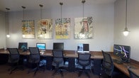This computer lab is a thoughtfully designed workspace that balances functionality with an engaging aesthetic. The room is outfitted with multiple workstations arranged along built-in countertop desks with a rich, dark granite-style surface, each station equipped with a desktop computer and ergonomic mesh chair. Pendant lighting suspended from the ceiling provides focused, warm illumination throughout the space, complementing the neutral gray walls and dark carpeted flooring.

A standout feature of the lab is the series of large, vibrant urban-themed art panels displayed prominently along the main wall, adding a creative and energetic character to what could otherwise be a utilitarian environment. The artwork features bold graphic illustrations of cityscapes, lending the room a dynamic and modern ambiance.

In addition to the primary row of workstations, the lab includes a dedicated printing station equipped with a laser printer, conveniently positioned near the entrance. A glass-paned door with a "B.Y.O.P. – Bring Your Own Paper" decal hints at a self-service print culture. A secondary seating area with additional computer terminals is also present, maximizing the lab's capacity. The overall layout is efficient and organized, making this space well-suited for academic, professional, or general-use computing needs.
