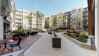 This thoughtfully designed outdoor courtyard serves as the central amenity hub of a contemporary multi-story residential community. Enclosed by a sweeping arc of modern, neutral-toned buildings with private balconies, the space creates a welcoming, resort-inspired atmosphere bathed in natural sunlight.

The courtyard is organized into several distinct zones, each designed for a specific purpose. A generously sized hardscape plaza, finished in light stone pavers, anchors the center and features a sleek, linear fire pit that serves as a focal point for gathering. Adjacent to it, a warm hardwood deck hosts outdoor dining tables and lounge seating, perfect for relaxing or entertaining. A covered pergola area with an intricate geometric lattice overhead filters sunlight into beautiful patterns across plush sectional sofas below.

Raised landscaped planters filled with drought-tolerant plants and agave add texture and greenery throughout, softening the hardscape transitions. A pool area with lounge chairs and market umbrellas is visible in the background, extending the recreational offerings.

Entryways featuring floor-to-ceiling glass storefronts and natural stone cladding provide elegant connections between the interior and exterior. Tall palm trees punctuate the perimeter, reinforcing the Southern California character of the development. The overall design balances functionality, aesthetics, and community-oriented living seamlessly.