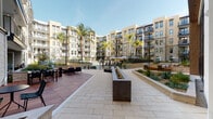 This thoughtfully designed outdoor courtyard serves as the central amenity hub of a contemporary multi-story residential community. Enclosed by a sweeping arc of modern, neutral-toned buildings with private balconies, the space creates a welcoming, resort-inspired atmosphere bathed in natural sunlight.

The courtyard is organized into several distinct zones, each designed for a specific purpose. A generously sized hardscape plaza, finished in light stone pavers, anchors the center and features a sleek, linear fire pit that serves as a focal point for gathering. Adjacent to it, a warm hardwood deck hosts outdoor dining tables and lounge seating, perfect for relaxing or entertaining. A covered pergola area with an intricate geometric lattice overhead filters sunlight into beautiful patterns across plush sectional sofas below.

Raised landscaped planters filled with drought-tolerant plants and agave add texture and greenery throughout, softening the hardscape transitions. A pool area with lounge chairs and market umbrellas is visible in the background, extending the recreational offerings.

Entryways featuring floor-to-ceiling glass storefronts and natural stone cladding provide elegant connections between the interior and exterior. Tall palm trees punctuate the perimeter, reinforcing the Southern California character of the development. The overall design balances functionality, aesthetics, and community-oriented living seamlessly.