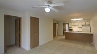 This single-floor unit offers an open-concept layout that seamlessly connects the living area to the kitchen, creating a spacious and functional flow throughout the interior. The living area features light-toned carpeting and neutral cream-colored walls, contributing to a bright and understated ambiance. A ceiling fan with an integrated light fixture is centrally mounted, providing both air circulation and overhead lighting to the main living space.

Warm-toned wooden doors are visible throughout, leading to additional rooms and hallways that branch off from the main open area. The kitchen is situated toward the rear of the space and is defined by medium-toned wood cabinetry, a light countertop surface, and a peninsula-style island that separates it from the living area. Appliances including a microwave and a gas range are integrated into the kitchen layout, and under-cabinet lighting adds functional illumination to the workspace.

The transition between the living area and kitchen is smooth, with consistent flooring tying the spaces together. The overall aesthetic is clean and practical, with simple finishes and a neutral palette that offers a versatile backdrop for a variety of interior styles.