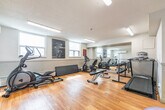 Located at 950 Warwick Crt in Burlington, Ontario, this fitness facility offers a well-equipped exercise space designed to support a range of workout routines. The room features warm-toned hardwood flooring throughout, white walls, and a textured ceiling fitted with overhead fluorescent lighting that keeps the space bright and functional.

A standout architectural feature is the large floor-to-ceiling mirror spanning one wall, which visually expands the room and provides users with proper form feedback during workouts. Natural light filters in through multiple windows, enhancing the open and energetic atmosphere of the space.

The facility is equipped with a variety of cardio and strength training equipment, including a treadmill, elliptical trainer, recumbent and upright stationary bikes, a rowing machine, and a dumbbell rack with a full range of free weights. A flat workout bench and exercise mat provide additional options for floor-based and strength exercises. Stability balls and a foam mat round out the amenities for flexibility and core training.

Climate control is provided by a wall-mounted split air conditioning unit, ensuring comfort year-round. Safety equipment, including a fire extinguisher, is visibly installed. The room is accessed through a glass-paneled door and is finished with clean, neutral décor accented by motivational fitness artwork on the walls.