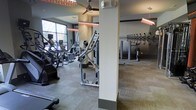 This contemporary fitness center is thoughtfully designed to provide a comprehensive workout environment. The space is organized into distinct exercise zones, accommodating both cardio and strength training needs. A generous selection of cardio equipment — including ellipticals, stair climbers, and other aerobic machines — lines the perimeter near large floor-to-ceiling windows that flood the room with natural light and offer views of the surrounding landscape. Structural columns divide the space, creating a sense of distinct workout areas while full-length wall mirrors enhance the sense of depth and allow exercisers to monitor their form. The strength and resistance training section features a variety of cable machines and weight equipment arranged for efficient use of the floor space. Overhead, warm-toned linear pendant lighting runs across the ceiling alongside a ceiling fan, ensuring the environment remains comfortable and well-lit throughout. The flooring consists of durable, neutral-toned carpet tiles, providing both cushioning underfoot and easy maintenance. The overall ambiance is clean, modern, and functional, with a neutral color palette of grays and whites that creates an energizing yet focused atmosphere suited for a full range of fitness activities.
