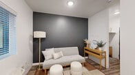 This single-floor unit offers a thoughtfully designed layout that maximizes functionality within a compact footprint. The kitchenette is a standout feature, outfitted with light wood-grain cabinetry that extends from floor to ceiling, providing generous storage space. Granite-patterned countertops offer ample prep surface, while the stainless single-basin sink, black refrigerator, built-in cooktop, and microwave ensure the space is fully equipped for everyday use. Warm wood-look plank flooring runs throughout, tying the interior together with a cohesive, contemporary feel. Natural light filters in through blinds-fitted windows, brightening the open kitchen and living area. A bold charcoal accent wall in the main room adds a striking visual contrast against the otherwise crisp white walls, lending the space a modern character. A wall-mounted HVAC unit provides independent climate control. The bathroom is clean and practical, featuring the same wood-look flooring, white walls, a toilet, a shower area, and grab bar details that suggest accessibility-conscious design. Overall, the unit presents a fresh, move-in-ready interior with a neutral palette and modern finishes suited for comfortable everyday living.
