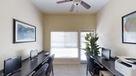 This single-floor space includes a well-appointed office area designed to accommodate multiple users simultaneously. The room features dual workstations positioned along opposing walls, each equipped with dark-finished desks and ergonomic seating, providing an efficient and organized workspace. Light tile flooring runs throughout, contributing to a clean, professional aesthetic.

Natural light enters generously through a large window fitted with horizontal blinds, allowing for adjustable brightness throughout the day. An adjacent glass-paneled door further enhances the sense of openness and provides additional light. The walls are finished in a neutral warm tone, creating a calm and focused atmosphere conducive to work.

A ceiling fan is centrally mounted, offering both practical air circulation and a subtle design element. A decorative indoor plant adds a touch of greenery, softening the functional nature of the space. A framed map or artwork on the wall provides a modest decorative accent. The overall ambiance is practical, clean, and comfortable, making this space well-suited for use as a home office or shared workspace.
