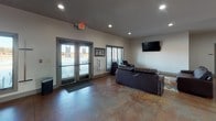 This single-floor facility is designed as a versatile communal space, offering a well-rounded mix of recreational, social, and functional areas unified by rich, polished concrete flooring that extends throughout the entire layout.

The main lounge area serves as the centerpiece of the space, furnished with leather sofas and armchairs arranged around a coffee table atop a decorative area rug. A wall-mounted flat-screen TV and unique sculptural wall art add character and comfort to this gathering zone. Recessed lighting provides consistent illumination across the open floor plan.

Adjacent to the lounge, a spacious game room houses a full-size pool table and additional seating, making it well-suited for recreational activities and social events. The open sightlines between these areas contribute to a cohesive and inviting atmosphere.

A curved bar with high-top seating and pendant lighting overhead creates a distinct social hub within the space. Framed artwork adorns the nearby walls, and a secondary service area is visible alongside the bar. Natural light enters through double-entry glass doors and surrounding windows, enhancing the bright and welcoming feel.

A dedicated office, accessible from the main hall, provides a private workspace with a desk and computer setup. The layout also includes additional corridors and doorways that suggest further utility rooms or support spaces, rounding out the functionality of this well-appointed facility.