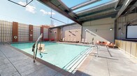 This impressive rooftop amenity space offers a comprehensive resort-style experience within an urban setting. At its center is a generously sized swimming pool with crystal-clear water, accessible via stainless steel handrails and equipped with an ADA-compliant pool lift. The pool is framed by a semi-covered structure with exposed steel beams and large skylight panels that flood the deck with natural light while providing partial shade. A row of lounge chairs lines the poolside, complemented by vibrant orange seating arrangements that create inviting social zones throughout the deck.

Adjacent to the pool area, a well-appointed outdoor kitchen and gathering space features multiple cooking islands with overhead ventilation hoods, bar-height seating, and pendant lighting suspended from the industrial-style ceiling. String lights add a warm ambiance to this entertaining zone. Plush, sectional lounge seating in bold red tones offers comfortable relaxation areas, perfect for socializing or unwinding. A dedicated screening room rounds out the amenities, featuring a large projection screen, colorful artwork, and cozy lounge chairs — ideal for private viewings or events. The overall design blends industrial architecture with warm, vibrant furnishings for a dynamic and functional communal experience.
