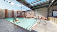 This impressive rooftop amenity space offers a comprehensive resort-style experience within an urban setting. At its center is a generously sized swimming pool with crystal-clear water, accessible via stainless steel handrails and equipped with an ADA-compliant pool lift. The pool is framed by a semi-covered structure with exposed steel beams and large skylight panels that flood the deck with natural light while providing partial shade. A row of lounge chairs lines the poolside, complemented by vibrant orange seating arrangements that create inviting social zones throughout the deck.

Adjacent to the pool area, a well-appointed outdoor kitchen and gathering space features multiple cooking islands with overhead ventilation hoods, bar-height seating, and pendant lighting suspended from the industrial-style ceiling. String lights add a warm ambiance to this entertaining zone. Plush, sectional lounge seating in bold red tones offers comfortable relaxation areas, perfect for socializing or unwinding. A dedicated screening room rounds out the amenities, featuring a large projection screen, colorful artwork, and cozy lounge chairs — ideal for private viewings or events. The overall design blends industrial architecture with warm, vibrant furnishings for a dynamic and functional communal experience.