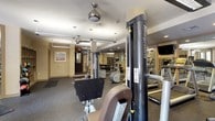 This fitness center is a fully outfitted, single-floor facility designed to support a wide range of workout routines. The space is thoughtfully organized into distinct zones, beginning with a dedicated cardio area lined with treadmills, elliptical machines, and stair climbers by Precor and similar brands. Wall-mounted flat-screen televisions are positioned for convenient viewing during cardio sessions. Adjacent to this area, a strength training zone features Magnum cable machines, free weights, dumbbells, and weight benches arranged on durable rubber flooring. Large mirrors line the walls throughout, enhancing the sense of space and allowing users to monitor their form. A separate stretching and functional training area is bathed in natural light from large windows overlooking an outdoor patio, and features stability balls in multiple sizes along with additional free weights neatly stored on racks. Ceiling fans provide additional air circulation throughout the main workout floor. The facility also includes access to restroom facilities via glass-paned interior doors, and framed artwork adds a welcoming touch to the neutral-toned walls. The overall ambiance is clean, bright, and well-maintained, making it a practical and comfortable environment for fitness enthusiasts of all levels.