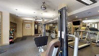 This fitness center is a fully outfitted, single-floor facility designed to support a wide range of workout routines. The space is thoughtfully organized into distinct zones, beginning with a dedicated cardio area lined with treadmills, elliptical machines, and stair climbers by Precor and similar brands. Wall-mounted flat-screen televisions are positioned for convenient viewing during cardio sessions. Adjacent to this area, a strength training zone features Magnum cable machines, free weights, dumbbells, and weight benches arranged on durable rubber flooring. Large mirrors line the walls throughout, enhancing the sense of space and allowing users to monitor their form. A separate stretching and functional training area is bathed in natural light from large windows overlooking an outdoor patio, and features stability balls in multiple sizes along with additional free weights neatly stored on racks. Ceiling fans provide additional air circulation throughout the main workout floor. The facility also includes access to restroom facilities via glass-paned interior doors, and framed artwork adds a welcoming touch to the neutral-toned walls. The overall ambiance is clean, bright, and well-maintained, making it a practical and comfortable environment for fitness enthusiasts of all levels.
