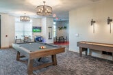 This generously proportioned single-floor community space is thoughtfully designed to accommodate socializing, dining, and relaxation. The open-concept layout seamlessly connects a living room, dining area, and fully equipped kitchen, creating a versatile environment for gatherings of all sizes.

The living room area anchors the space with a cozy fireplace, plush seating including a mix of sofas and accent chairs, and large glass entry doors that invite abundant natural light and outdoor views. Elegant drum-style chandeliers illuminate the room, complementing the patterned carpeting underfoot.

A standout feature is the full bar and kitchen area, highlighted by a dramatic natural stone accent wall that serves as a striking backdrop for dual wall-mounted televisions. The expansive white quartz island with bright blue metal barstools provides ample seating, while warm wood cabinetry, stainless steel under-counter appliances, and draft beer taps complete a premium entertainment setup.

The dining areas are distributed throughout, offering both casual bistro-height tables near the entrance and intimate low-top seating accented by a vibrant red area rug. A curated gallery wall featuring eclectic framed artwork and mirrors adds personality and warmth to the neutral gray walls. A central billiard table serves as a focal point, making this a well-rounded space designed for community engagement.