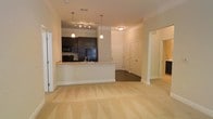 This single-floor residence is designed around an open-concept floor plan that seamlessly connects the kitchen and living areas, creating a cohesive and functional living environment. The kitchen is a standout feature, showcasing dark espresso-toned cabinetry that contrasts sharply with the light-colored peninsula countertop. The peninsula includes an undermount sink and provides a natural dividing point between the kitchen and the main living area. Black appliances, including a refrigerator, range, and built-in microwave, complement the dark cabinetry aesthetic. Overhead, pendant lights hang from the ceiling above the peninsula, while a track lighting system illuminates the kitchen workspace, adding both style and practicality.

The living area is finished with plush, light-toned carpeting that extends throughout, contributing to a warm and comfortable atmosphere. Neutral wall tones and crisp white baseboards run throughout the space, maintaining a clean and cohesive visual style. Beyond the kitchen, a hallway leads to additional areas of the unit, with paneled doors and archways visible in the background, hinting at further private spaces. The overall ambiance is tidy, modern, and move-in ready.