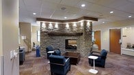 This single-floor space makes a strong first impression through its carefully designed lounge area, anchored by a commanding stacked-stone fireplace that stretches toward the ceiling. The fireplace surround, constructed of layered natural stone in earthy tones of gray and brown, is topped with a substantial wood mantel and accented by recessed spotlights that highlight the texture and craftsmanship of the stonework. A dark wood header frames the curved upper portion of the fireplace feature wall, adding architectural definition and visual contrast.

The seating area is arranged thoughtfully around the fireplace, with navy upholstered armchairs and small accent tables providing a comfortable and functional gathering space. A central dark wood coffee table grounds the arrangement. The patterned carpet in warm, neutral tones adds visual interest underfoot and contributes to the overall inviting atmosphere.

Overhead, a dropped ceiling with a suspended tile grid incorporates recessed lighting throughout, ensuring the space is well-lit. The combination of warm materials, ambient lighting, and a functional fireplace gives the space a professional yet welcoming character well-suited for communal use.