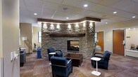 This single-floor space makes a strong first impression through its carefully designed lounge area, anchored by a commanding stacked-stone fireplace that stretches toward the ceiling. The fireplace surround, constructed of layered natural stone in earthy tones of gray and brown, is topped with a substantial wood mantel and accented by recessed spotlights that highlight the texture and craftsmanship of the stonework. A dark wood header frames the curved upper portion of the fireplace feature wall, adding architectural definition and visual contrast.

The seating area is arranged thoughtfully around the fireplace, with navy upholstered armchairs and small accent tables providing a comfortable and functional gathering space. A central dark wood coffee table grounds the arrangement. The patterned carpet in warm, neutral tones adds visual interest underfoot and contributes to the overall inviting atmosphere.

Overhead, a dropped ceiling with a suspended tile grid incorporates recessed lighting throughout, ensuring the space is well-lit. The combination of warm materials, ambient lighting, and a functional fireplace gives the space a professional yet welcoming character well-suited for communal use.