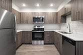 This thoughtfully designed one-bedroom, one-bathroom unit offers a comfortable and efficient layout across a single floor. The combined kitchen and living area serves as the heart of the space, featuring an L-shaped kitchen outfitted with stainless steel appliances — including a refrigerator, electric range, built-in microwave, and dishwasher. Sleek wood-toned cabinetry, white quartz countertops, a classic subway tile backsplash, and light luxury vinyl plank flooring give the kitchen a clean, modern aesthetic. The living area includes a wall-mounted mini-split HVAC unit and a glass-panel door that allows natural light to filter in. Neutral gray walls and recessed lighting are consistent throughout the main living spaces. The bedroom is carpeted for comfort and includes a window with blinds providing natural light, a walk-in closet with built-in shelving, and direct hallway access to the bathroom. The bathroom features a wood-toned vanity with a white countertop, a large mirror, and a full shower-tub combo. A dedicated laundry room houses a full-size front-loading washer and dryer, adding everyday convenience. Light, neutral finishes unify the space with a cohesive, contemporary feel.