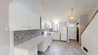 Located at 31 Putnam Green in Greenwich, CT, this two-story 1-bedroom, 1-bathroom residence offers a thoughtfully designed layout spread across two levels. The kitchen serves as a standout feature, showcasing bright white cabinetry extending to the ceiling for generous storage, a striking black-and-white mosaic penny tile backsplash, light-toned countertops, and wood-look tile flooring. A window above the sink brings in natural light, and a pendant light fixture accents the entry area near the front door, which features a classic sunburst transom window. The upper level includes a spacious bedroom with plush carpeting, two large closets with warm wood-toned doors, a window with outdoor views, and a wall-mounted HVAC unit alongside baseboard heating. The full bathroom presents a wide white vanity with an oversized mirror and clean, neutral finishes. The lower level offers an open carpeted living area with a textured ceiling, staircase access, and additional door storage. Throughout, the neutral gray and white palette creates a clean, cohesive aesthetic suited for comfortable everyday living in the heart of Greenwich.