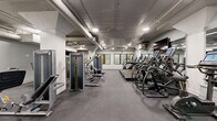 Located at 2400 Grayland Ave in Richmond, Virginia, this fitness center offers a comprehensively outfitted exercise space across a single, generously sized floor. The facility is designed with a functional, industrial aesthetic, featuring exposed ductwork and overhead piping along the concrete ceiling, complemented by recessed lighting that keeps the space well-illuminated throughout.

The main exercise area is expansive, providing ample room for a wide array of workout equipment. A full row of treadmills and elliptical machines lines one side, offering dedicated space for cardio training. Opposite, a robust selection of strength machines, including cable and weight-stack systems, are thoughtfully arranged to maximize floor space and ease of use. A well-stocked free weight section features a comprehensive dumbbell rack alongside additional resistance equipment, catering to a variety of training preferences.

Large wall-mounted mirrors are positioned throughout the main floor, enhancing the sense of space while allowing users to monitor their form. A flat-screen television is mounted prominently for entertainment during workouts.

A separate, enclosed studio room adjoins the main floor, finished in lighter tones with its own mirrored walls, light-colored flooring, and glass-paneled doors. This studio is equipped with stability balls, resistance equipment, and yoga mats, making it ideal for stretching, group classes, or personal training sessions. The durable rubber flooring throughout ensures safety and comfort across all workout zones.