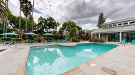 Located at 2949 Portage Bay W in Davis, California, this outdoor recreational amenity area serves as a central gathering space within a well-established residential community. The centerpiece is a generously sized swimming pool with crystal-clear blue water, accessible via built-in entry steps and pool-side railings. Adjacent to the main pool sits a separate hot tub spa, offering a dedicated space for relaxation. The surrounding pool deck is finished in a smooth concrete surface and is furnished with teal lounge chairs and patio seating sets shaded by matching teal umbrellas, creating a cohesive and inviting aesthetic. The space is framed by mature trees — including tall palm trees and broad deciduous varieties — alongside lush, manicured shrubs and plantings that provide privacy and a natural, park-like atmosphere. The residential buildings visible in the background feature a contemporary design with wood-panel siding and clean angular lines, contributing to the community's cohesive architectural character. A clubhouse or amenity building with large sliding glass doors and a covered overhang adjoins the pool area, offering convenient indoor-outdoor access. The overall ambiance is relaxed and resort-like, designed to encourage outdoor leisure and community interaction in a verdant Northern California setting.