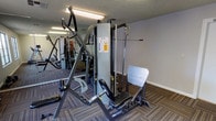 This fitness center is a single-floor space designed to support a full range of strength training and exercise activities. The room is outfitted with professional-grade cable and weight machines, including a multi-function strength station with adjustable seats and cable pulley systems, catering to a variety of muscle groups and workout routines. Mirrored walls line one side of the space, visually expanding the room while allowing users to monitor their form during exercise. The flooring consists of durable, striped commercial carpeting that provides comfort and stability underfoot. Overhead fluorescent lighting ensures the space is well-lit throughout. The walls are finished in a neutral gray tone with a textured ceiling, giving the room a clean and functional aesthetic. The layout is thoughtfully arranged to allow users to move between equipment with ease. Overall, this fitness center offers a practical and well-maintained environment equipped to meet the needs of regular gym-goers and fitness enthusiasts alike.