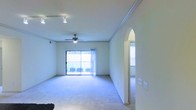 This single-floor space presents a well-proportioned open living area designed with both functionality and comfort in mind. The room is finished with soft carpeted flooring throughout, providing a warm underfoot feel. Crown molding runs along the perimeter of the ceiling, adding a classic architectural detail that enhances the overall character of the space.

Overhead, a ceiling fan with an integrated light fixture serves as a central focal point, while a track lighting system along one wall offers additional, directional illumination. Natural light floods the interior through a wide sliding glass door, which opens onto an outdoor balcony with a railing—bringing in views of greenery beyond and creating a seamless connection between interior and exterior.

A distinctive arched doorway on one side of the living area leads to an adjoining room, which also benefits from natural light through a nearby window. This architectural arch adds a subtle design accent that breaks from the otherwise clean, neutral aesthetic of the space. The walls are finished in a light, neutral tone, contributing to an open and airy ambiance throughout.