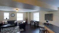 This single-floor space offers a thoughtfully designed open-concept layout that seamlessly blends living and dining areas into one cohesive environment. The interior is finished with rich, dark hardwood floors that add warmth and contrast against the light, neutral-toned walls painted in soft yellow and gray tones. A ceiling fan with integrated lighting provides both airflow and ambient illumination, complementing the natural light that streams in through multiple windows fitted with white blinds. The living area is anchored by a pair of sleek dark leather armchairs arranged atop a bold geometric patterned area rug, creating an inviting and stylish seating arrangement. A decorative wall clock and ornamental vase add personality to the space. Adjacent to the seating area, a compact dining or workspace setup features a dark wood table with matching chairs, positioned near a window to take advantage of natural light. A wall-mounted decorative panel adds an artistic accent to the room. Toward the edge of the frame, a counter-height surface with a metal bar stool hints at an additional casual dining or kitchen bar area, suggesting a functional and versatile floor plan suited to everyday living.