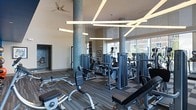 This fitness center offers a thoughtfully designed workout environment characterized by its impressive ceiling height, which lends the space an open, airy feel. Striking linear pendant lights intersect overhead in a geometric pattern, complementing the recessed lighting and a ceiling fan that enhances air circulation throughout the room. Tall, mosaic-tiled cylindrical columns add a decorative architectural element, while large floor-to-ceiling windows along one wall invite abundant natural light and provide views of the surrounding exterior. The flooring features a combination of patterned carpet tiles in neutral tones, adding texture and visual interest underfoot. The equipment layout is generous and well-organized, with a comprehensive selection of strength-training machines, cable stations, weight benches, and rowing equipment distributed throughout the space. A dedicated wall niche adds a subtle design accent near the entrance area. The overall ambiance is contemporary and polished, balancing functionality with refined aesthetics to create an inviting and motivating fitness environment suitable for a wide range of exercise routines.