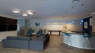 This single-floor space offers a well-appointed communal lounge designed for both casual relaxation and social interaction. The open-plan layout is anchored by warm wood-look flooring and a neutral color palette accented by soft teal and blue tones throughout.

The seating area features plush gray sectional sofas and teal accent chairs arranged to encourage conversation, complemented by a sleek white side table. Nearby, a high-top curved bar counter with chrome barstools serves as a central gathering point, illuminated by striking cobalt blue pendant lights that add a bold design touch.

A shuffleboard table occupies the middle of the room, providing an entertaining focal point for recreational use. Additional high-top seating with slender bar chairs can be found along the perimeter, offering flexible arrangements for smaller groups.

The walls are adorned with colorful framed artwork that adds visual interest and a contemporary feel to the space. Recessed lighting and flush-mount ceiling fixtures ensure the room is well-lit throughout. Subtle architectural details such as tray ceiling transitions and clean crown molding contribute to the polished, modern aesthetic. Overall, the space strikes a balance between inviting comfort and stylish functionality.