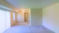 This single-floor residence offers a straightforward and functional layout designed for comfortable everyday living. The main living area is finished with wall-to-wall carpet in a neutral tone, providing a soft and cohesive foundation throughout the space. Walls are painted in a light, warm beige that helps reflect natural light and creates an open, airy atmosphere. A ceiling fan with an integrated light fixture is centrally positioned, ensuring both comfort and adequate illumination. A window along one wall allows natural light to filter into the room, adding warmth and brightness to the interior. The layout flows naturally from the main living area through an open passageway into a connecting hallway, which leads to additional rooms beyond, including what appears to be a bedroom or secondary space with a door visible in the background. The hallway provides a clear sense of connectivity throughout the unit, making navigation between spaces seamless. Neutral wall colors and consistent flooring throughout contribute to a clean, uniform aesthetic. The space is move-in ready and offers a blank canvas well-suited for personalization. Overall, this residence provides a practical and welcoming environment with a calm, understated design sensibility.