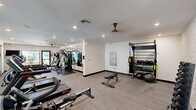 Located at 20707 N 67th Ave in Glendale, Arizona, this fitness center offers a thoughtfully designed exercise space ideal for a well-rounded workout experience. The room features a clean, contemporary aesthetic with neutral-toned walls, textured stone accent walls, and durable low-pile carpeted flooring throughout. Recessed lighting and ceiling fans ensure the space remains bright and comfortable during use.

The cardio section is well-appointed with treadmills, elliptical trainers, stationary bikes, and rowing machines, providing ample options for aerobic exercise. A dedicated strength training area includes a multi-station cable machine, adjustable weight benches, a full rack of dumbbells, and a functional training rig equipped with TRX suspension straps, resistance accessories, and medicine balls.

Large floor-to-ceiling glass walls and sliding glass doors connect the gym to an adjacent lounge area, flooding the space with natural light and creating a sense of openness. Additional windows along the exterior walls offer views of the surrounding outdoor greenery, enhancing the overall ambiance. Wall-mounted televisions are positioned for convenient viewing during cardio sessions. The space is efficiently organized to maximize usability while maintaining an open, uncluttered feel.
