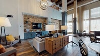 This thoughtfully designed single-floor communal space blends modern sophistication with warm, inviting elements throughout its open-concept layout. The main lounge area commands attention with dramatic soaring ceilings accented by exposed wood beam detailing and an eye-catching orb chandelier that casts warm light across the room. A striking textured stone mosaic accent wall serves as the focal point, framing a large flat-screen television and flanked by slate-blue built-in cabinetry with open shelving — perfect for both display and storage. Rich hardwood-style flooring anchors the space and flows seamlessly through the lounge and into adjoining areas. Mid-century modern furnishings, including a walnut credenza, paired table lamps, and plush seating, lend a curated, residential feel. A dedicated kitchenette area features natural wood cabinetry, white hexagonal tile flooring, a wall-mounted television, and ample counter space with a undermount sink. An adjacent bar-height island with stylish pendant lighting and upholstered counter stools provides an ideal space for casual gathering or work. Additional seating nooks and a well-stocked bookshelf area offer versatile options for relaxation. Large windows throughout invite natural light, enhancing the warm, welcoming ambiance of the entire space.