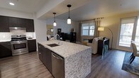 This thoughtfully designed single-floor space showcases a contemporary open-concept layout that effortlessly connects the kitchen, dining, and living areas into one cohesive living environment. The kitchen serves as the centerpiece, anchored by a generous island topped with striking light-toned granite countertops featuring bold veining. Dark cabinetry provides a sharp contrast to the stone surfaces, while stainless steel appliances — including a built-in dishwasher — add a sleek, modern touch. Pendant lighting with clean, cylindrical shades hangs above the island, casting warm, focused illumination over the workspace. The flooring throughout is a warm-toned wood-look material that lends a sense of continuity and warmth across the entire space. The open living area beyond the kitchen is filled with natural light streaming through large windows dressed with neutral curtains, creating an airy and inviting ambiance. A comfortable seating arrangement and a media console define the living zone, while the dining area sits adjacent, completing the open-plan flow. The overall aesthetic blends modern design with warm, residential comfort — making this a functional and visually appealing space suited for everyday living and entertaining alike.