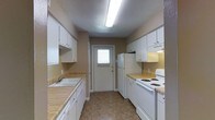 This single-floor space is designed with practicality and functionality at its core. The kitchen is laid out in a classic galley style, with countertops and cabinetry lining both sides of a central walkway, maximizing the use of available space. White shaker-style cabinets extend from base to upper levels on both walls, offering generous storage throughout. The countertops feature a warm laminate finish that complements the neutral, tan-toned walls and textured ceiling. On one side, a white double-basin sink sits beneath upper cabinetry, with additional counter space ideal for food preparation. The opposite wall houses a white electric coil range and oven, accompanied by an over-the-range exhaust hood and upper cabinets. A full-size white refrigerator anchors the end of one counter run. The flooring is a durable, earth-toned vinyl tile that ties the space together with a clean, low-maintenance finish. At the far end of the kitchen, a single door with a small window allows for natural light to filter in, brightening the space alongside the overhead fluorescent lighting. The overall ambiance is clean, utilitarian, and well-organized, suited for efficient day-to-day use.