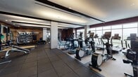 Located at 710 12th St in Arlington, VA, this premium fitness center is thoughtfully designed to accommodate a full range of workout needs across multiple distinct training zones. The space features a sophisticated aesthetic, with warm wood-paneled walls, recessed and pendant lighting, and a refined dark color palette that lends a sleek, modern feel throughout.

The cardio zone is positioned along expansive floor-to-ceiling windows, flooding the area with natural light and offering open views of the Arlington skyline. A row of high-tech treadmills and cycling stations are situated here, complemented by stylish circular pendant fixtures overhead.

The free weights area is lined with large mirrors that enhance the sense of space, and houses a fully stocked dumbbell rack alongside benches and cable machines. Rubberized flooring in this section provides durability and safety for heavy lifting.

The functional training zone features a wooden plyometric platform, a heavy tire, barbell racks with weight plates, and a wall-mounted flat-screen television — catering to strength and performance-focused workouts.

A dedicated strength machine zone rounds out the facility, offering a variety of selectorized cable and resistance machines set on carpet-tiled flooring, with medicine balls and additional accessories available. The overall ambiance is that of a high-end, well-equipped fitness facility designed for serious training.