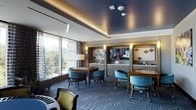 This single-floor amenity space is designed as an upscale game and entertainment lounge, blending sophisticated design with functional leisure. The room is anchored by a large round game table surrounded by yellow upholstered chairs, ideal for poker or card games, while teal barrel chairs arranged around smaller side tables offer additional seating areas for casual socializing or board games.

A striking navy blue tray ceiling adds depth and elegance to the space, complemented by warm recessed and cove lighting that creates an inviting, lounge-like ambiance. Patterned carpet in deep charcoal tones grounds the room and ties together the rich color palette throughout.

A large flat-screen television is mounted within a built-in cabinetry wall, flanked by decorative wall panels and soft sconce lighting. A wet bar or kitchenette runs along one wall, providing convenient service capability for gatherings. Floor-to-ceiling windows along one side of the room flood the space with natural light and offer expansive views of the surrounding cityscape. Glass-paneled doors provide access to an adjacent room, enhancing the overall openness and flow of the space.