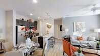 This 2-bedroom, 2-bathroom single-floor residence offers an open-concept design that integrates the kitchen, dining, and living areas into one cohesive, flowing space. The kitchen is appointed with dark cabinetry, stainless steel appliances, and light stone countertops, anchored by a central island with bar seating — ideal for casual dining or entertaining. Pendant lighting above the island adds a refined touch, complementing the recessed lighting throughout. The dining area accommodates a round glass table, positioned just off the kitchen for convenient access. The living area extends naturally from the dining space, featuring neutral-toned walls and light wood-style flooring that runs consistently throughout, lending warmth and visual continuity to the home. The layout feels spacious and well-proportioned, with an unobstructed sightline from the kitchen to the far end of the living room. The overall palette of soft grays and warm whites, accented by contemporary fixtures, creates a clean, modern aesthetic. The single-floor layout ensures easy navigation between all areas of the residence.