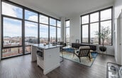 This single-floor unit is defined by its striking floor-to-ceiling windows framed in dark metal, which flood every room with natural light and offer expansive, elevated views of the surrounding cityscape. The open-concept living and dining area is anchored by the dramatic window wall, creating a seamless connection between the interior and the urban panorama beyond. Wide-plank hardwood flooring runs throughout the main living space, lending warmth to the otherwise contemporary aesthetic.

The kitchen is outfitted with high-gloss white cabinetry, stainless steel appliances including a gas range and french-door refrigerator, a subway tile backsplash, and a peninsula that provides additional counter space and casual seating. The layout flows naturally into the living area, making it well-suited for entertaining.

Both bedrooms benefit from large black-framed windows that continue the city views and allow generous natural light. The bathrooms feature neutral tile flooring, clean white cabinetry, extended vanities with under-mount sinks, and a glass-enclosed walk-in shower, all contributing to a polished, minimalist atmosphere throughout.