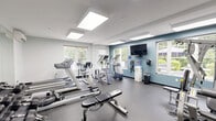Located at 95 Filley, this fitness center offers a thoughtfully designed exercise space laid out across a single floor. The room is bright and inviting, featuring large windows on multiple walls that flood the space with natural light and offer views of the surrounding greenery. Flush-mounted panel lighting on the white ceiling further enhances the open, airy atmosphere.

The exercise room is designed for a full-body workout experience. Cardio equipment includes multiple treadmills, an elliptical trainer, a stationary bike, and a WaterRower rowing machine. Strength training is well-represented with a multi-station cable machine, an adjustable weight bench, and a tiered dumbbell rack stocked with a wide range of free weights. Stability balls add flexibility training options to the mix.

A wall-mounted flat-screen television is positioned for easy viewing during workouts, and a water dispenser ensures convenient hydration. Full-length mirrors along one wall amplify the sense of space and allow for form checks during exercise. The durable speckled rubber flooring is practical and easy to maintain. A mini-split air conditioning unit provides climate control year-round. A glass partition and door connect the fitness room to an adjacent lounge area, adding a sense of community to the facility.