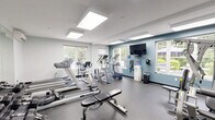 Located at 95 Filley, this fitness center offers a thoughtfully designed exercise space laid out across a single floor. The room is bright and inviting, featuring large windows on multiple walls that flood the space with natural light and offer views of the surrounding greenery. Flush-mounted panel lighting on the white ceiling further enhances the open, airy atmosphere.

The exercise room is designed for a full-body workout experience. Cardio equipment includes multiple treadmills, an elliptical trainer, a stationary bike, and a WaterRower rowing machine. Strength training is well-represented with a multi-station cable machine, an adjustable weight bench, and a tiered dumbbell rack stocked with a wide range of free weights. Stability balls add flexibility training options to the mix.

A wall-mounted flat-screen television is positioned for easy viewing during workouts, and a water dispenser ensures convenient hydration. Full-length mirrors along one wall amplify the sense of space and allow for form checks during exercise. The durable speckled rubber flooring is practical and easy to maintain. A mini-split air conditioning unit provides climate control year-round. A glass partition and door connect the fitness room to an adjacent lounge area, adding a sense of community to the facility.