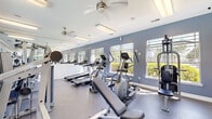 This fitness center offers a comprehensive workout environment thoughtfully designed to accommodate a range of exercise routines. The space is finished with a cool blue-gray color palette on the walls, white crown molding, and durable rubber flooring, creating a clean and functional aesthetic throughout.

Cardio equipment, including multiple treadmills and elliptical trainers, is arranged along one side of the room, providing ample space for users to exercise comfortably. Strength training is well represented with a selection of selectorized cable machines and weight machines positioned throughout the floor. A dedicated free weight area features a fully stocked dumbbell rack, accompanied by a flat bench for versatile lifting options.

Natural light pours in through a series of large windows fitted with horizontal blinds, offering pleasant views of the surrounding greenery. Ceiling fans are mounted above, promoting airflow and comfort during workouts. Full-length mirrors span the length of one wall, visually expanding the room and allowing users to monitor their form.

Overhead fluorescent lighting ensures the space is bright and well-lit at all times. A glass entry door connects the fitness center to the outdoor area, further enhancing the sense of openness. Overall, this fitness center is a well-appointed facility that balances functionality with a bright, inviting atmosphere.