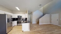 This two-story home is designed with a clean, modern aesthetic carried consistently throughout both levels. The main floor centers around an open-concept kitchen and living area, where light wood-tone flooring provides a warm, cohesive foundation. The kitchen is well-appointed with white shaker-style cabinetry, dark granite-look countertops, a black subway tile backsplash, and a full suite of stainless steel appliances including a gas range, microwave, and French door refrigerator. A peninsula with an undermount sink adds functional workspace and separates the kitchen from the adjacent living area, which features multiple windows and a glass door that invite ample natural light and outdoor views. A staircase with a metal railing leads to the upper level, where a spacious open loft area offers flexible use potential. The upper floor continues the light wood flooring and neutral color palette. Bedrooms are well-sized, each featuring ceiling fans, natural light from exterior windows, and closet space—including a walk-in closet with wrap-around hanging rods. Bathrooms echo the kitchen's design language with white vanities and dark countertops.