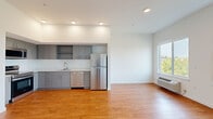This single-floor unit presents a clean, contemporary design throughout. The open-concept kitchen is well-appointed with a full suite of stainless steel appliances, including a refrigerator, electric range, over-the-range microwave, and dishwasher. Gray flat-front cabinetry with brushed hardware lines the walls, complemented by white quartz countertops and a stainless steel sink. The kitchen flows naturally into the open living area, which is finished with warm hardwood-style flooring that extends throughout the unit. Recessed lighting illuminates the space, while a wall-mounted PTAC unit provides climate control. Large windows in the living and sleeping areas fill the rooms with natural light and offer views of surrounding greenery. The bedroom areas feature tall ceilings and generous window placement, creating a bright, airy atmosphere. The bathroom is finished with gray cabinetry, a white quartz vanity top, an undermount sink, a large wall mirror, and a full tub-shower combination. Consistent use of neutral tones, modern fixtures, and durable finishes gives the unit a cohesive, move-in-ready aesthetic.