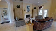 This single-story space showcases a thoughtfully designed living area that blends classic elegance with comfortable functionality. The room centers around a white marble fireplace, which serves as the focal point and is flanked by a pair of matching antique-white armoire cabinets featuring geometric relief detailing. A framed landscape painting is displayed prominently above the mantle, enhancing the room's refined aesthetic. Wall sconces on either side of the fireplace add a warm, ambient glow to complement the recessed lighting overhead.

The flooring consists of light-toned tile, contributing to the room's bright and airy feel. Crown molding lines the ceiling, adding an architectural detail that reinforces the classical design sensibility. Natural light filters into the space through a window dressed with rich burgundy valances and sheer curtains, offering a pleasant connection to the outdoors.

The seating arrangement includes upholstered armchairs in muted tones and a boldly patterned sofa with tropical leaf motifs, providing visual contrast. A round wooden side table at the center displays tasteful decorative accents, including ceramic vessels and a decorative orb, completing the polished, welcoming atmosphere of this living space.