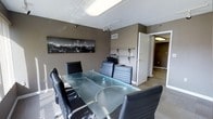 This business center presents a well-appointed, single-floor professional environment designed to support productive work and seamless collaboration. The centerpiece of the space is a stylish conference room featuring a large glass-top table surrounded by ergonomic black leather executive chairs, creating a sleek and contemporary atmosphere ideal for meetings and presentations. The walls are finished in a warm taupe tone, lending the space a grounded, professional character. Track lighting and recessed ceiling fixtures provide ample, even illumination throughout. A striking black-and-white cityscape photograph adorns the wall, adding a sophisticated urban touch to the decor. Built-in wall shelving offers practical storage and display space, complemented by a printer station that keeps office essentials neatly organized. The flooring is covered in neutral carpet that ties the room together cohesively. A corridor visible from the conference area suggests additional rooms or workspaces beyond, hinting at further functional areas within the center. Overall, this business center strikes a balance between modern design and practical utility, making it a polished and professional setting suited for a variety of business needs.