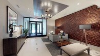 The lobby at 1177 Warburton Ave in Yonkers, NY presents a refined and welcoming first impression through its thoughtfully curated design. The entrance is framed by floor-to-ceiling glass doors that invite natural light deep into the space, complemented by large glass-panel walls that maintain an open, airy feel throughout.

A dramatic woven wood and gold textured accent wall serves as the lobby's centerpiece, adding warmth, depth, and visual interest to the neutral, light-toned palette. Light porcelain tile flooring with subtle geometric inlay patterns runs throughout, providing a clean and polished foundation. Recessed lighting and a statement ring chandelier illuminate the space with a warm, layered glow.

The seating area is elegantly appointed with contemporary upholstered chairs and a sofa, arranged around a glass-and-wood coffee table atop a geometric-patterned area rug. Decorative elements — including sculptural accents, potted greenery, abstract framed artwork, and a geometric-patterned credenza — add personality and sophistication.

A long corridor extends from the lobby, lined with smooth wall finishes and patterned carpeting, providing seamless access to the building's interior. A glass-enclosed lounge area is visible from the main lobby, reinforcing the sense of transparency and connected, modern design. The overall ambiance is polished, upscale, and inviting.